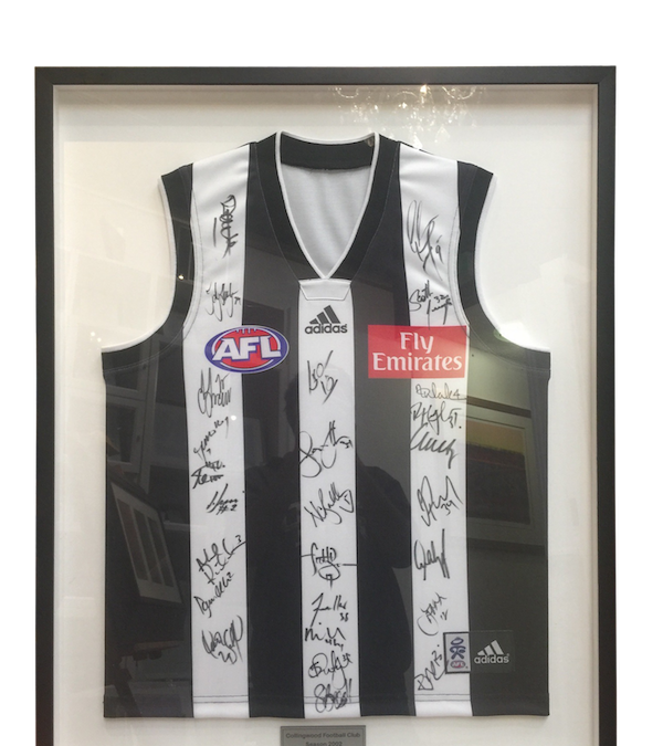 This is a magpies footy jersey and plaque signed by the entire team framed in a black box frame with the jersey set back from the UV glass on a mat board.