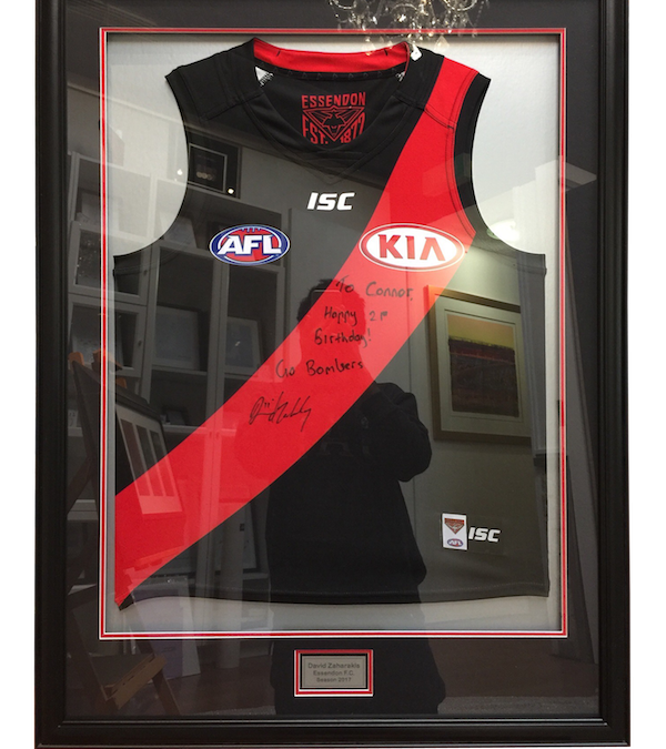 An Essendon Football Club jersey signed by David Zaharakis framed in a black frame with a double mat board surround.