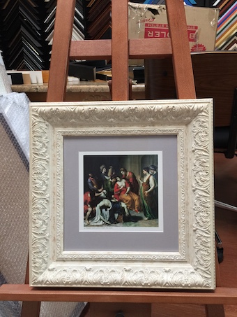A small antique print framed in a wide white ornate timber frame with a mat board surround and museum glass.