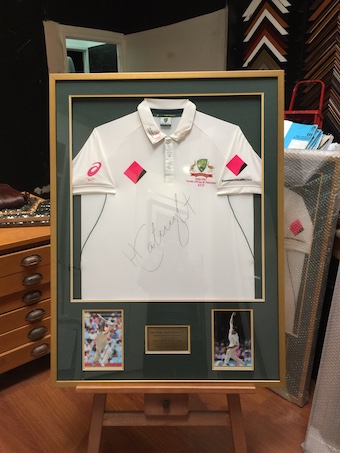 A cricket jersey framed in a gold box frame with multi opening mat board surround, photos and gold plaque.
