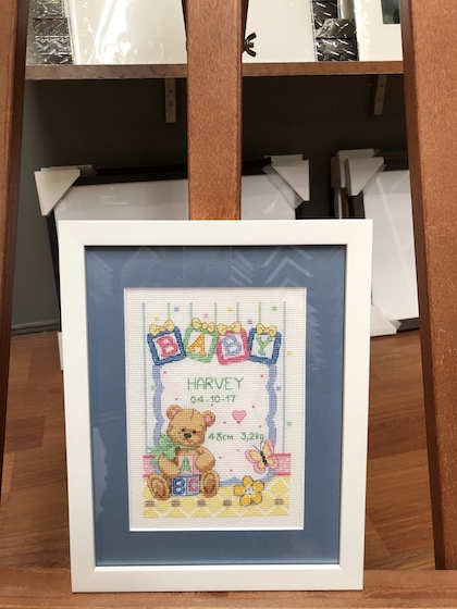 A childrens needlework framed in a white square frame with a mat board and glass.