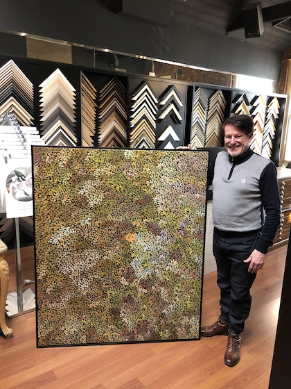 Greg Mahoney with a large Aboriginal painting stretched and framed in a black floater frame.