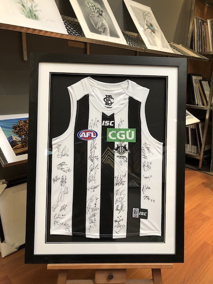 A Collingwood footy jumper signed by the team framed in a wide black box frame with double mat board surround.
