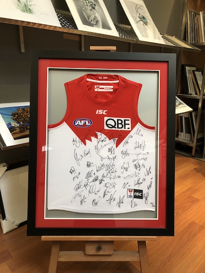 A Sydney Swans footy jumper hand signed by the team framed in a wide black timber frame with double mat board surround and glass.
