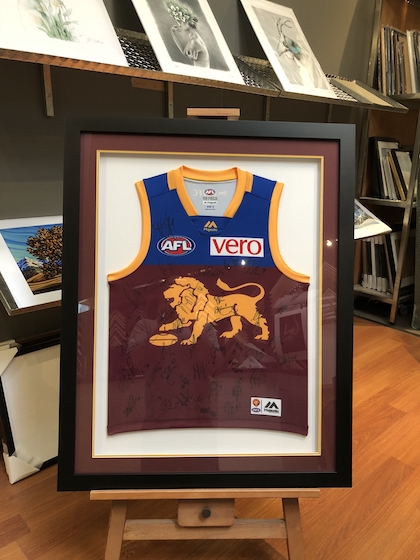 A Brisbane Lions footy jumper hand signed by the team framed in a black timber frame with double mat board surround and glass front.