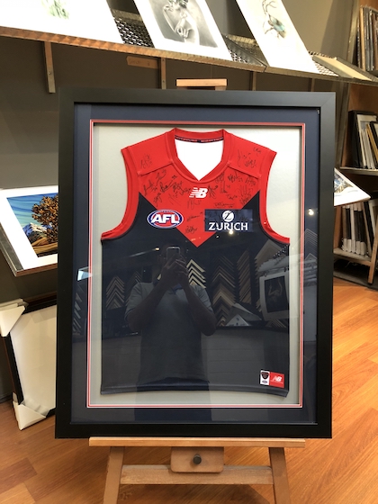 A Melbourne Demons footy jumper framed in a black timber frame with double mat boards and glass front.