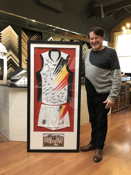 A sports jersey, shorts and photo framed in a large black timber frame with double mat boards and glass front.